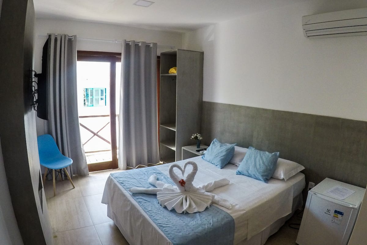 Quarto de hotel com cama, uma cadeira, guarda-roupa e mesa com abajur.