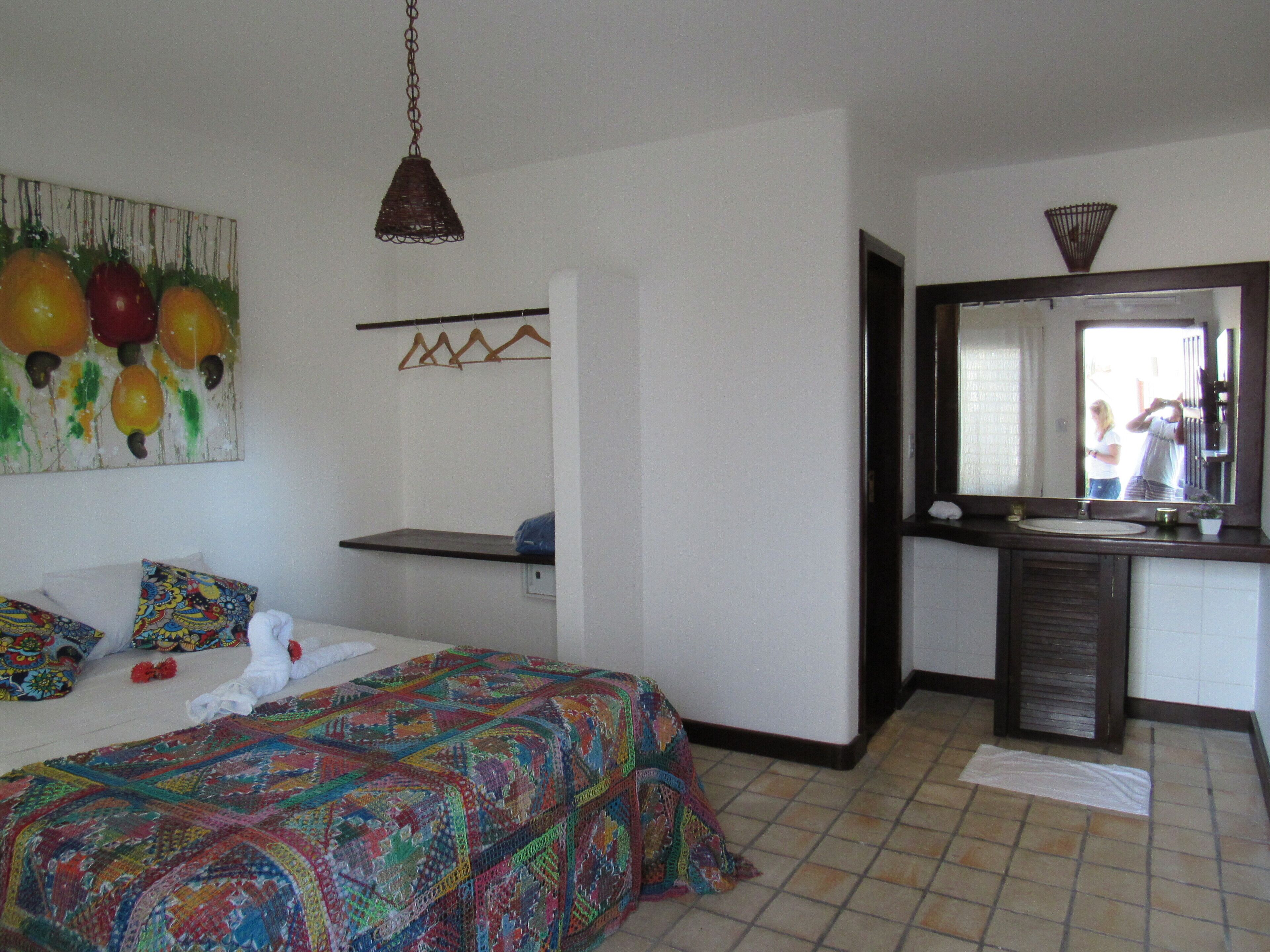 Un dormitorio con cama, colcha colorida, lámpara colgante, baño con espejo y un cuadro de frutas en la pared.