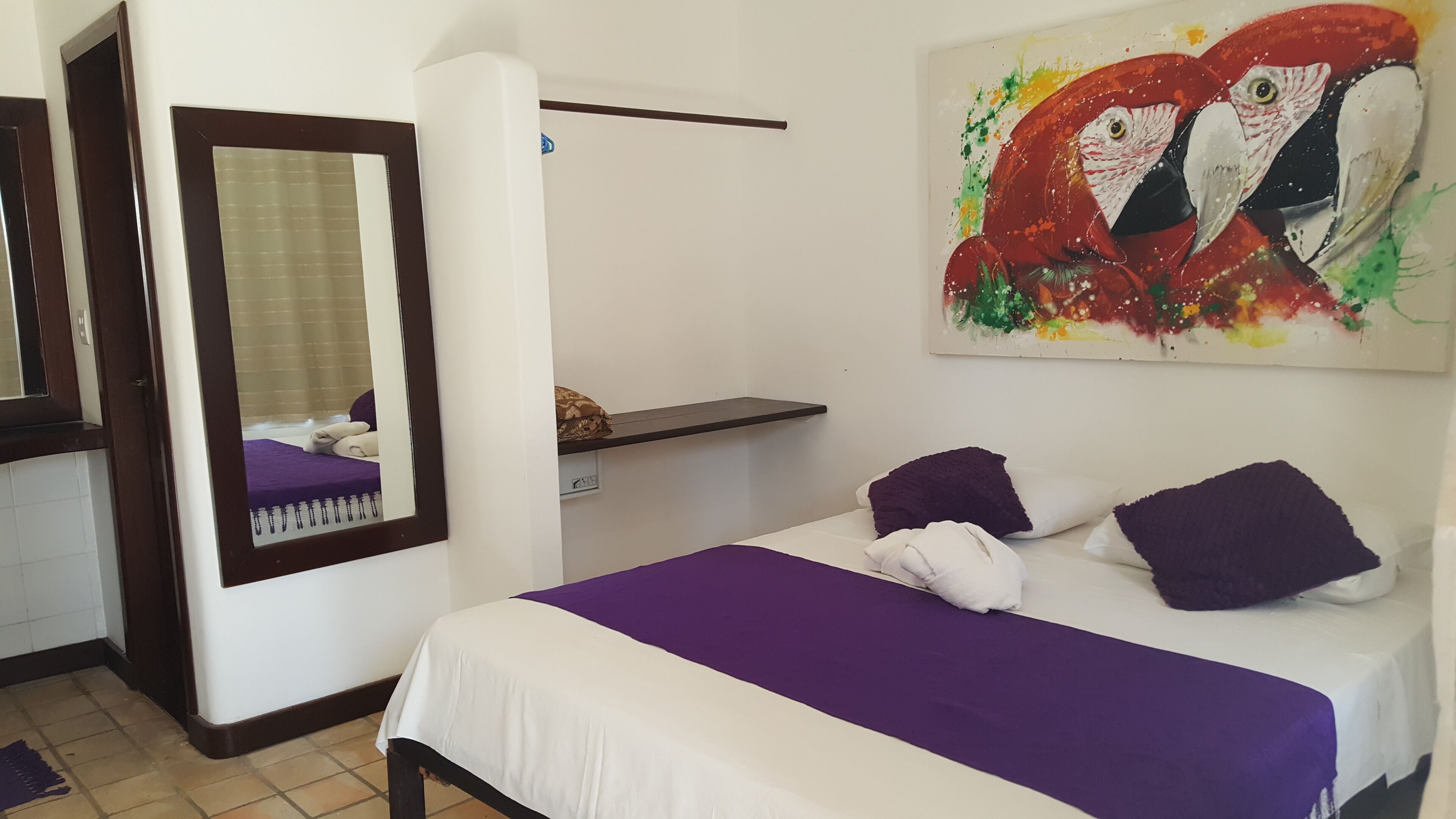 A bedroom with a bed, a mirror, a shelf, and a colorful painting.