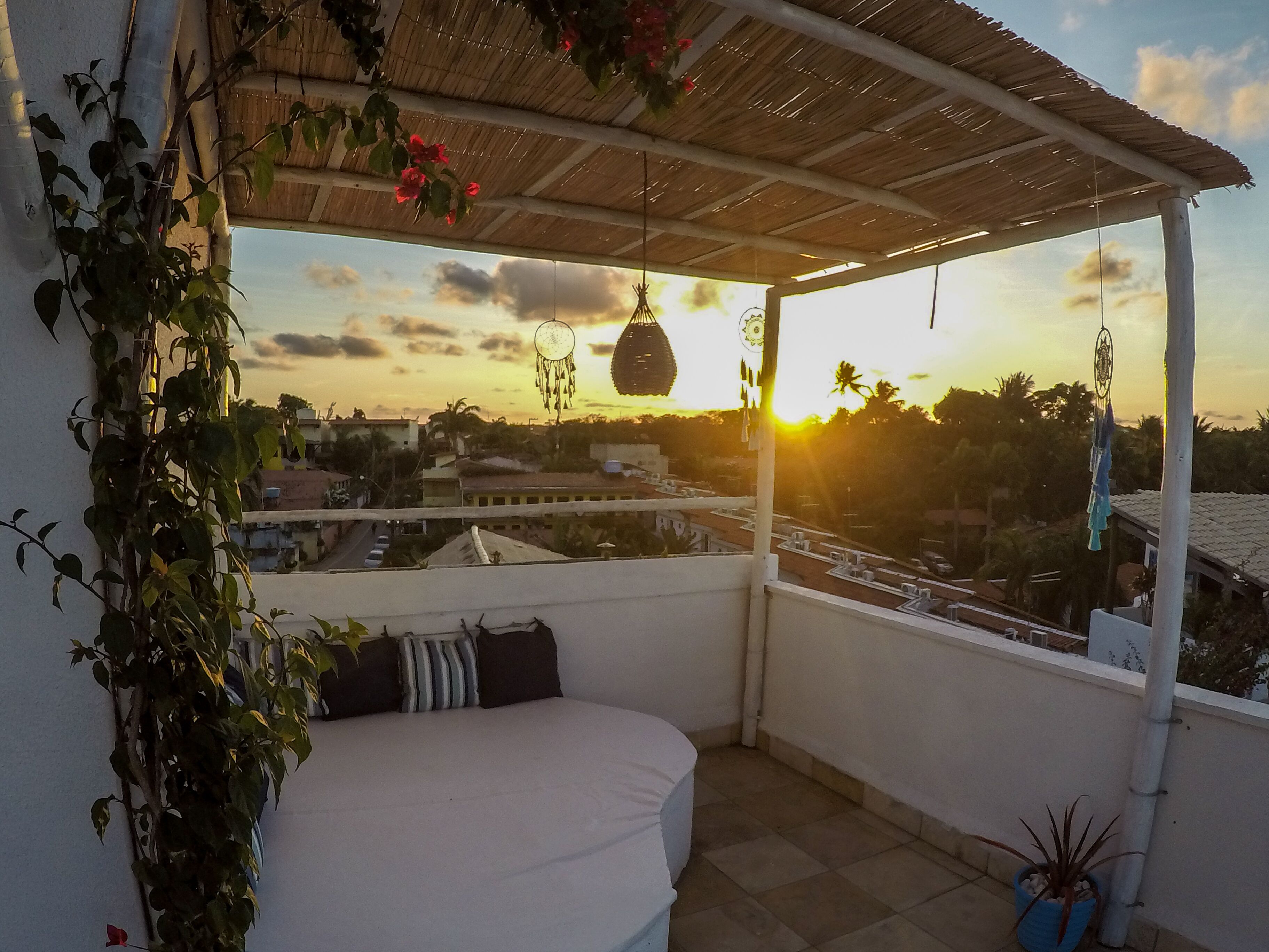 A balcony with a white cushioned seating area, a hanging dreamcatcher, and a view of the sunset over a cityscape.