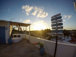 Terraza en la azotea con vista al atardecer, un panel con nombres de destinos, una puerta azul y plantas en macetas.