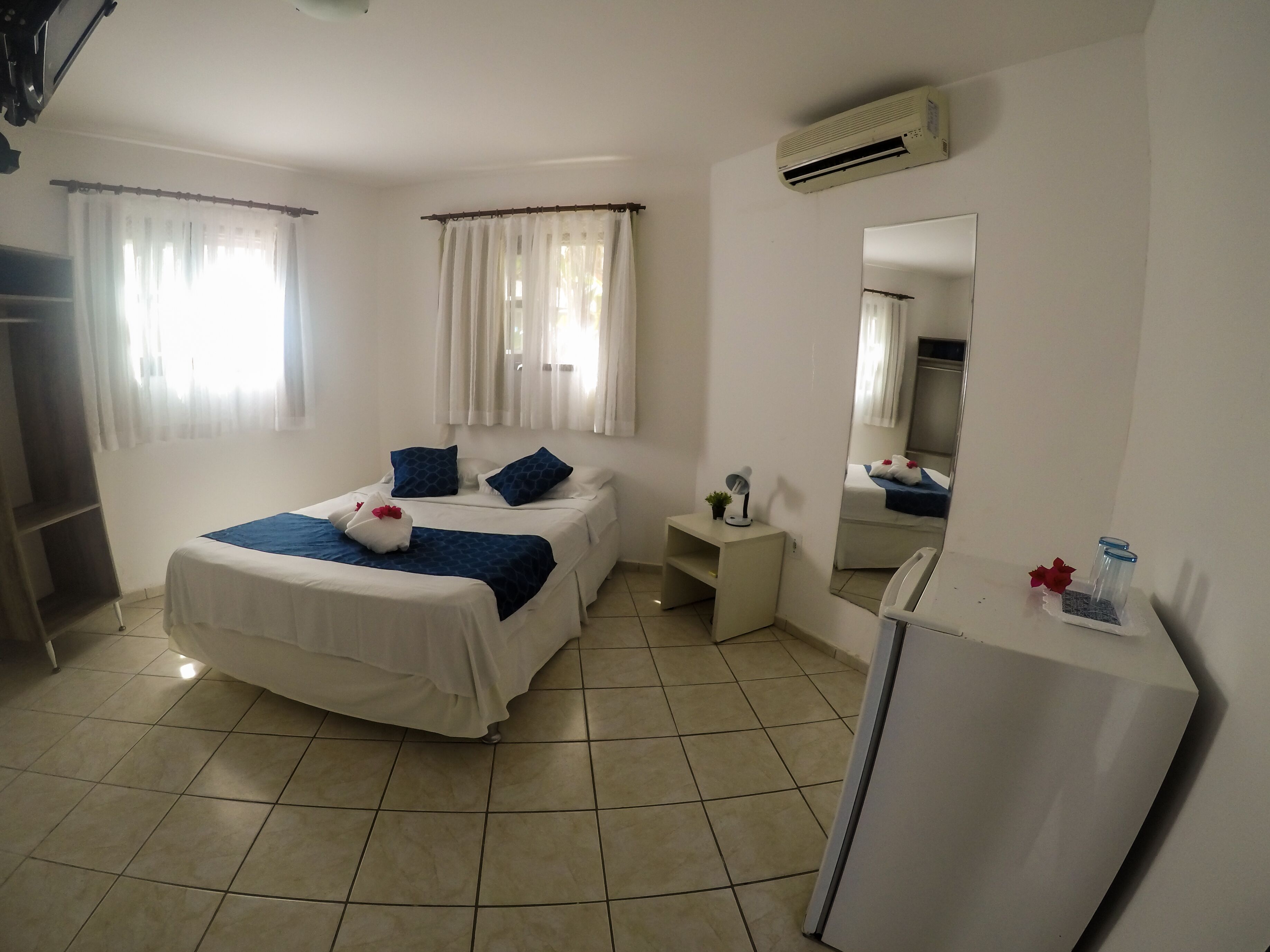 A hotel room with a bed, a small desk, a mirror, and an air conditioner.