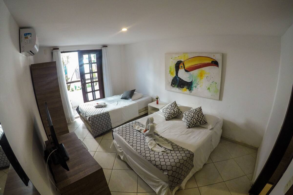 Quarto de hotel com duas camas, uma janela grande, um quadro de tucano e uma escadaria.