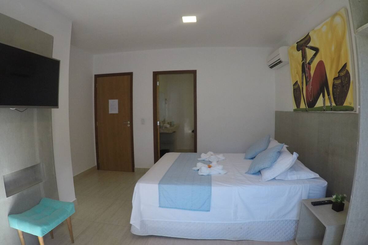 Quarto de hotel com cama, uma mesinha, uma cadeira, televisão presa na parede e um quadro na parede.