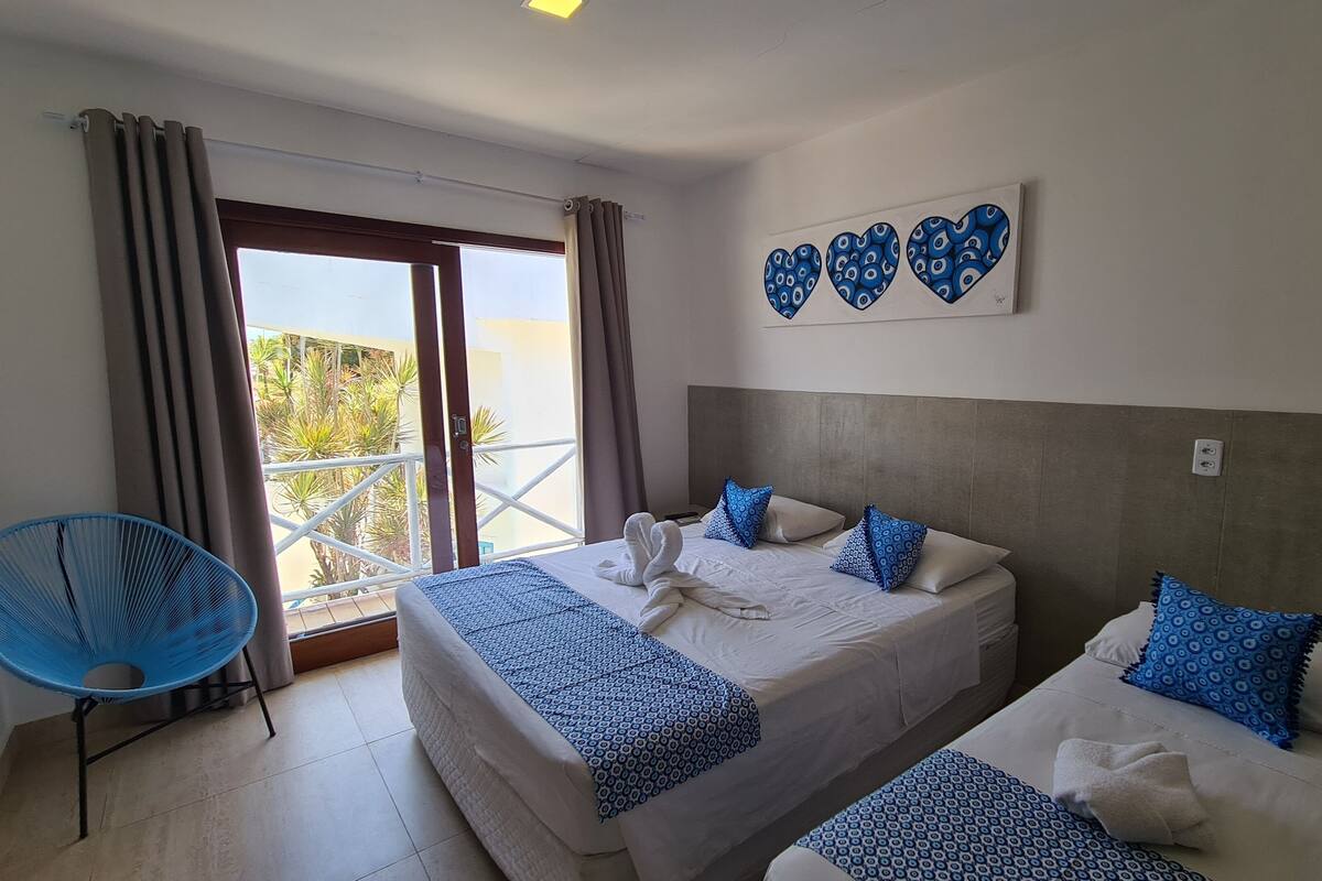 Quarto de hotel com duas camas, uma cadeira azul e uma varanda com cortinas.