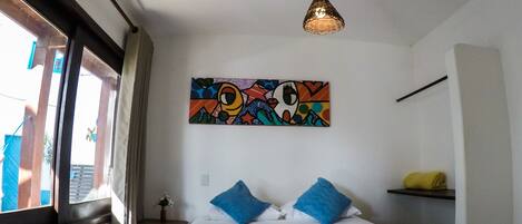 Ein Hotelzimmer mit Bett, einem farbenfrohen Kunstwerk an der Wand, einer Pendelleuchte und einem Fenster mit Vorhängen.