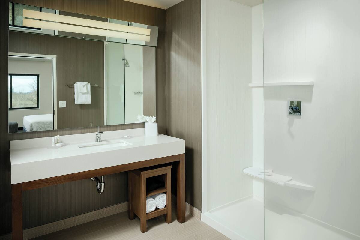 A modern bathroom with a large mirror, a double sink vanity, a wooden shelf with towels, and a separate shower area.