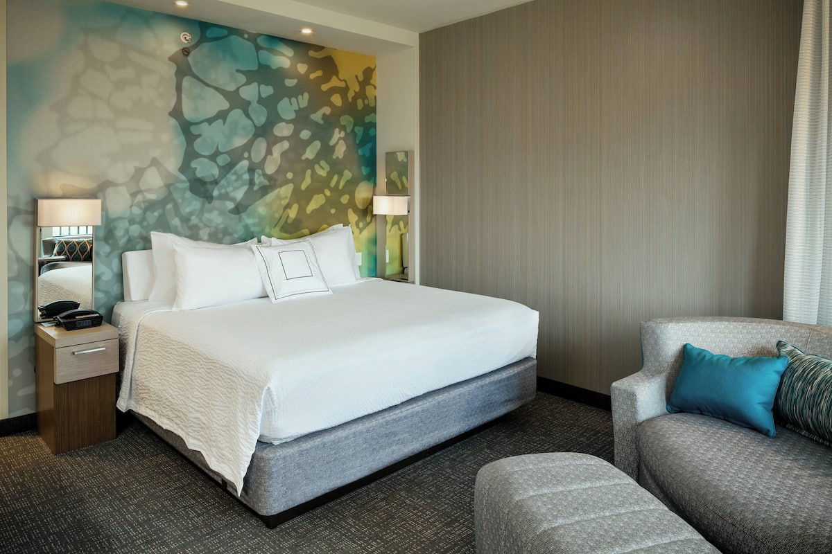 A hotel room with a large bed, a gray armchair, a nightstand with a lamp, and a wall with a colorful abstract design.