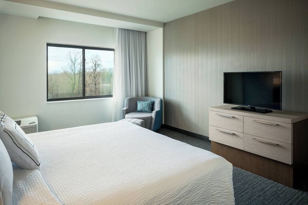 A modern hotel room with a large bed, a TV on a wooden cabinet, a chair with a pillow, and a window with curtains.