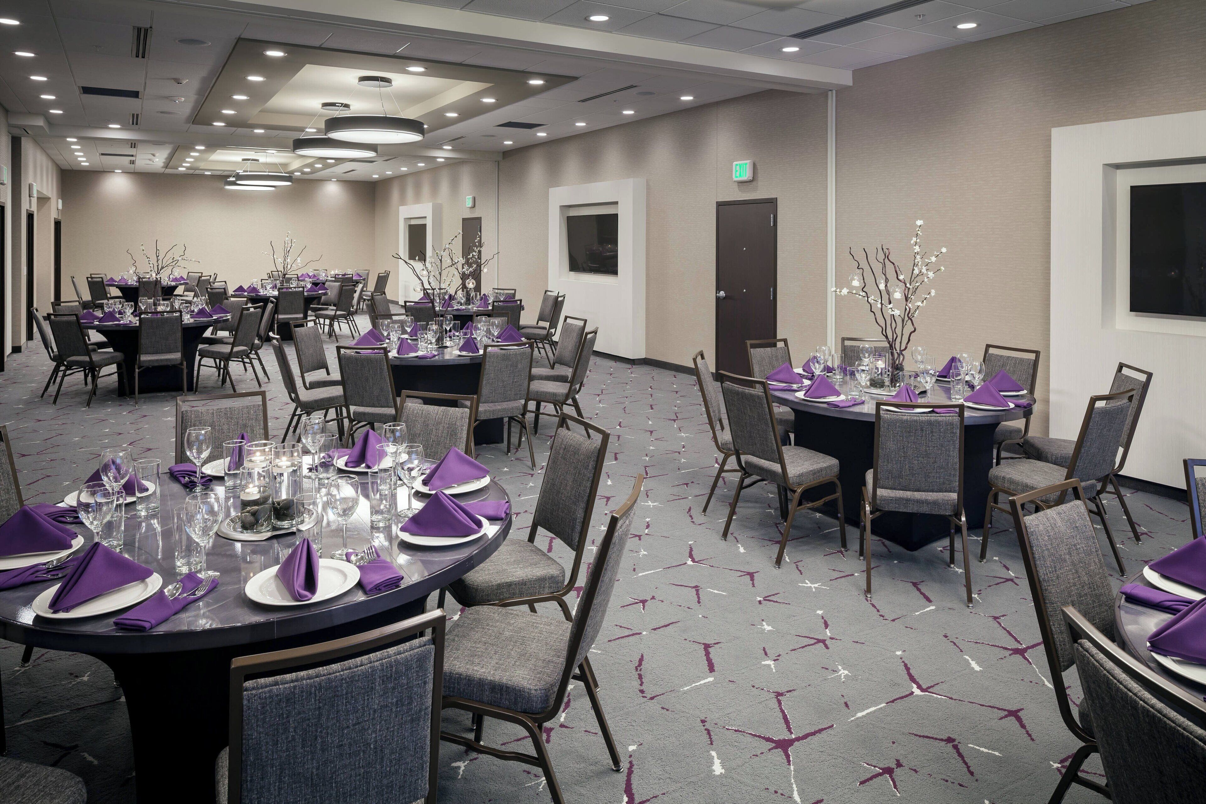 Une salle de conférence d’hôtel avec plusieurs tables rondes, nappes violettes et verres, chaises grises à revêtement en tissu et un tapis à motifs.