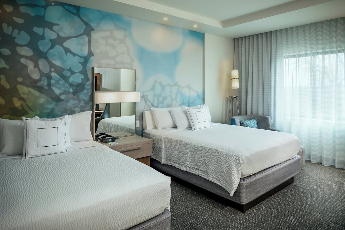 A hotel room with two beds, a nightstand, a lamp, and a large window with curtains.
