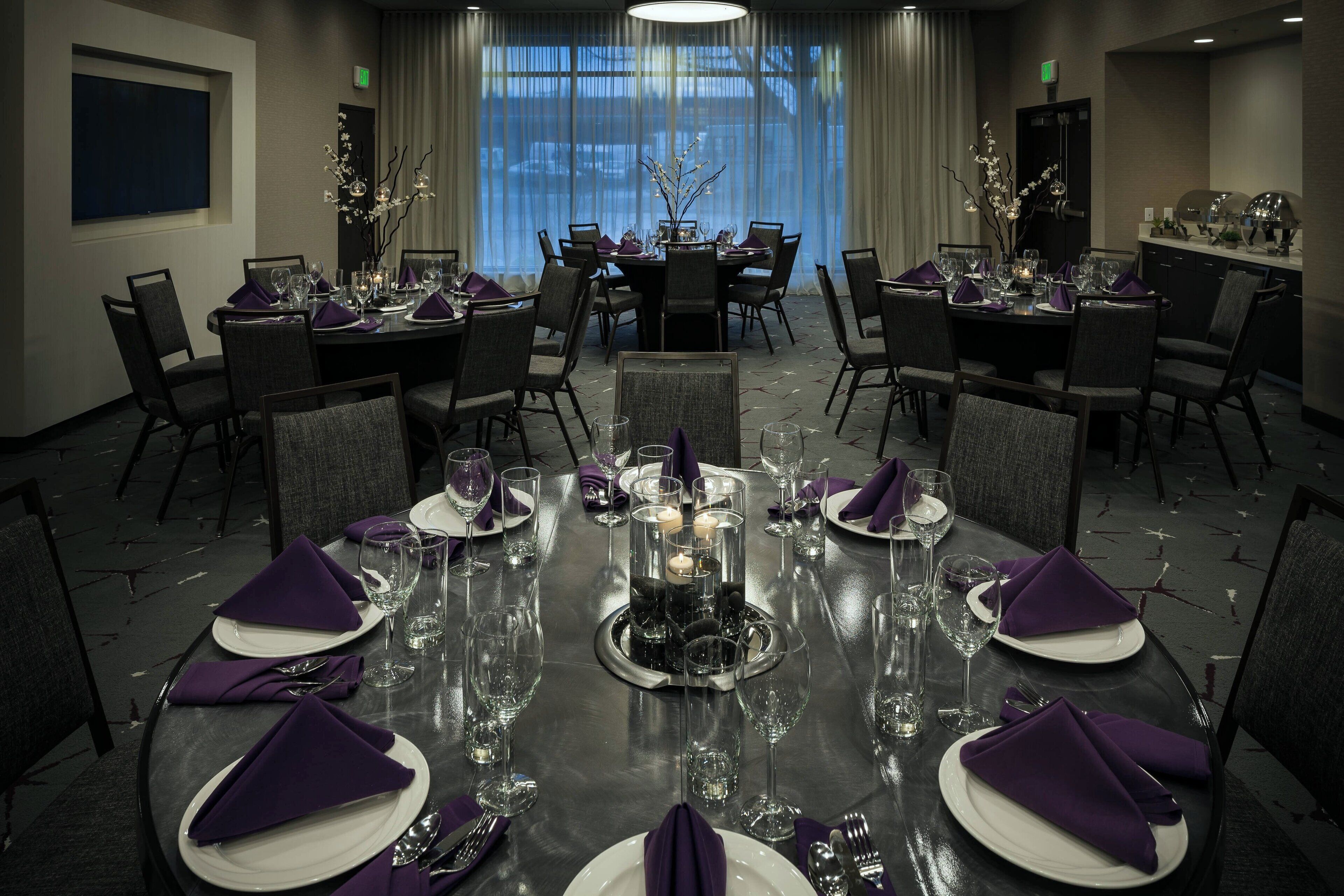 A dining area with round tables set for four, each with white plates, purple napkins, and clear glasses.