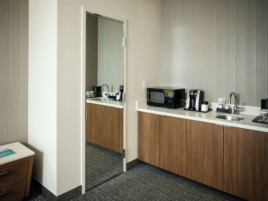 A modern hotel bathroom with a wooden vanity, a sink, a mirror, a microwave, and a coffee maker.