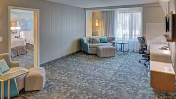 Habitación de hotel con sofá, sillón, reposapiés, escritorio y televisor.