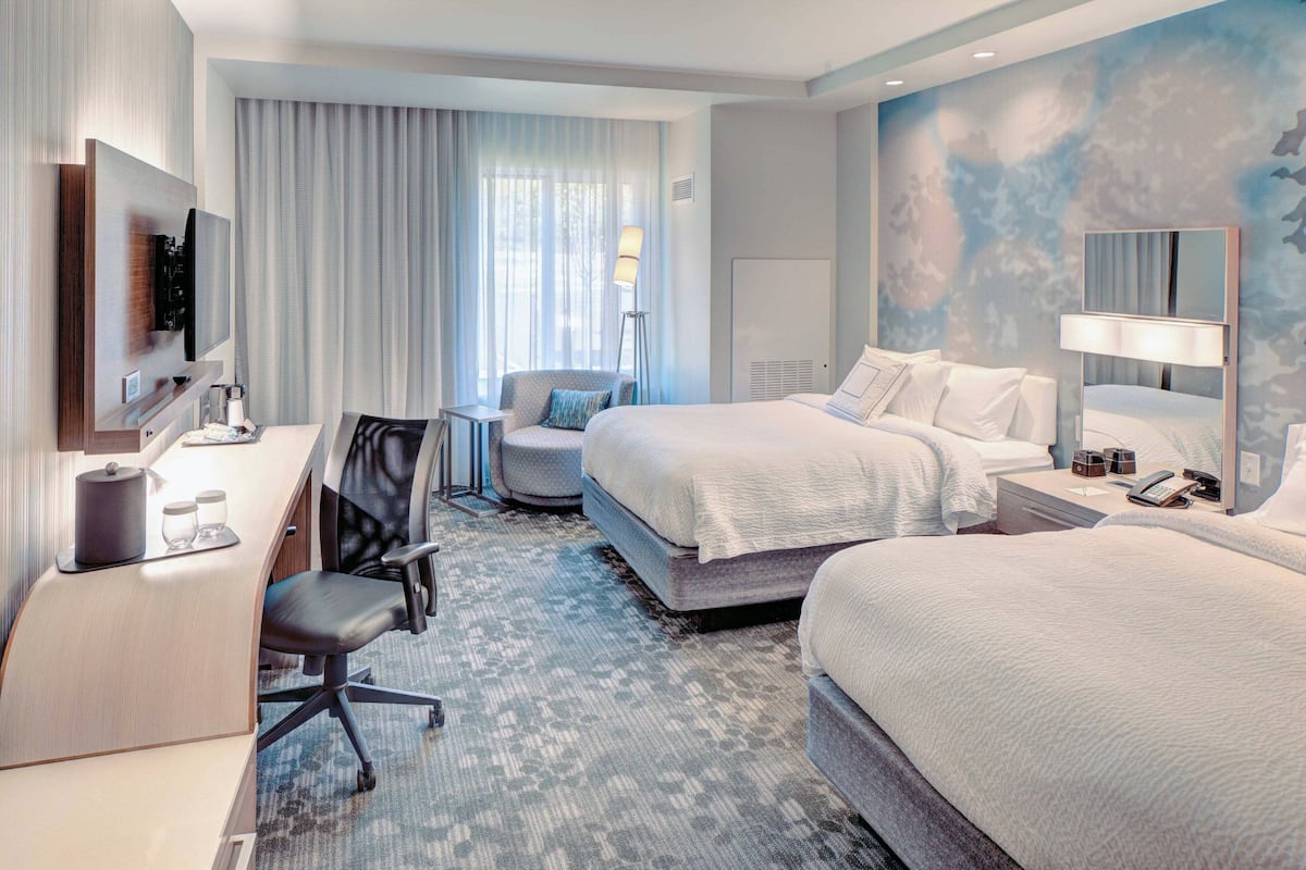 Une chambre d’hôtel équipée d’un bureau, de deux lits, d’une chaise, d’une lampe et d’une grande fenêtre avec des rideaux.