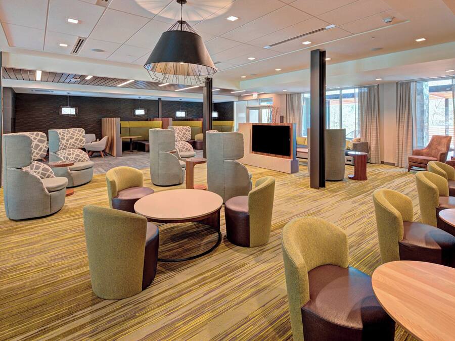 Eine moderne Hotelhalle mit verschiedenen Sitzgelegenheiten, darunter Sofas und Sessel, die um eine zentrale Fläche mit einem Flachbildfernseher angeordnet sind.