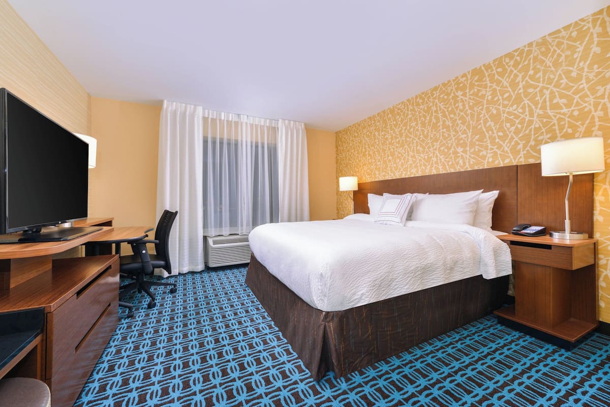 A hotel room with a bed, a desk with a television, a chair, a bedside table with a lamp, and a patterned carpet.