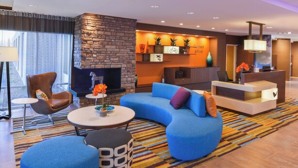 Una sala de estar moderna con un sofá azul, un sillón marrón y una mesa de centro redonda. Incluye una chimenea, un mostrador de recepción y una alfombra colorida a rayas.