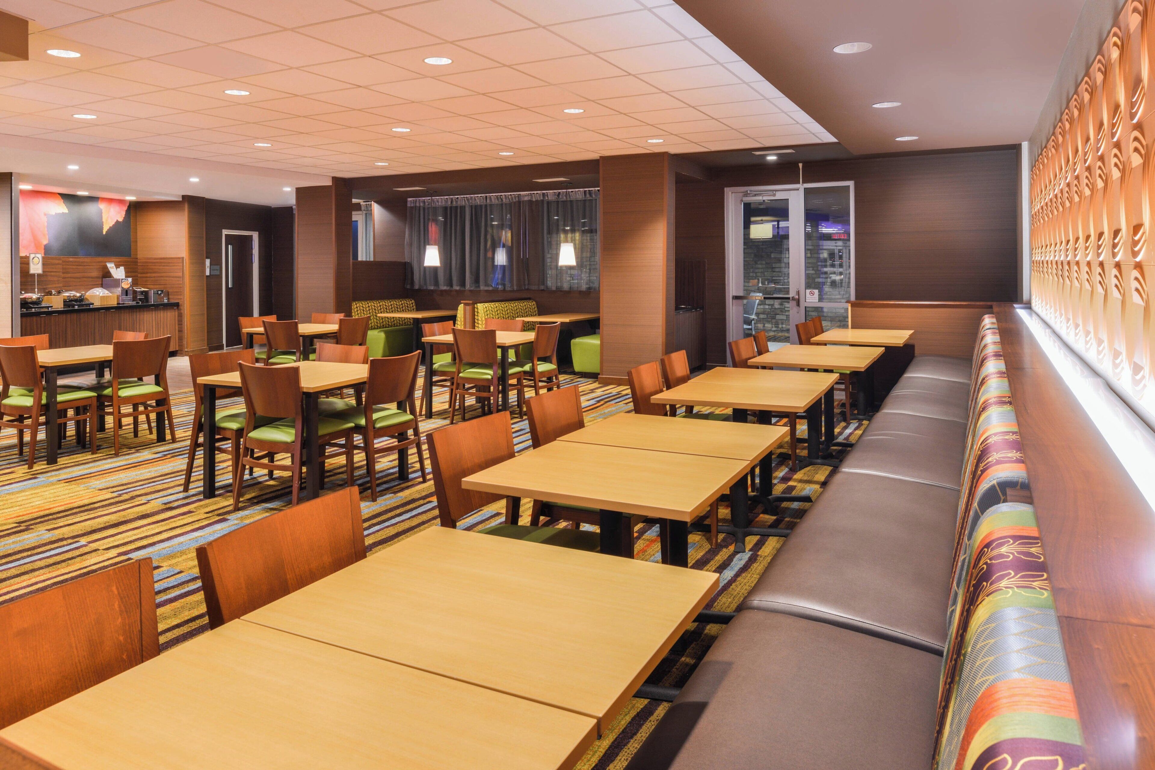 A hotel dining area with multiple tables and booths, carpeted flooring, and a wall with a patterned design.