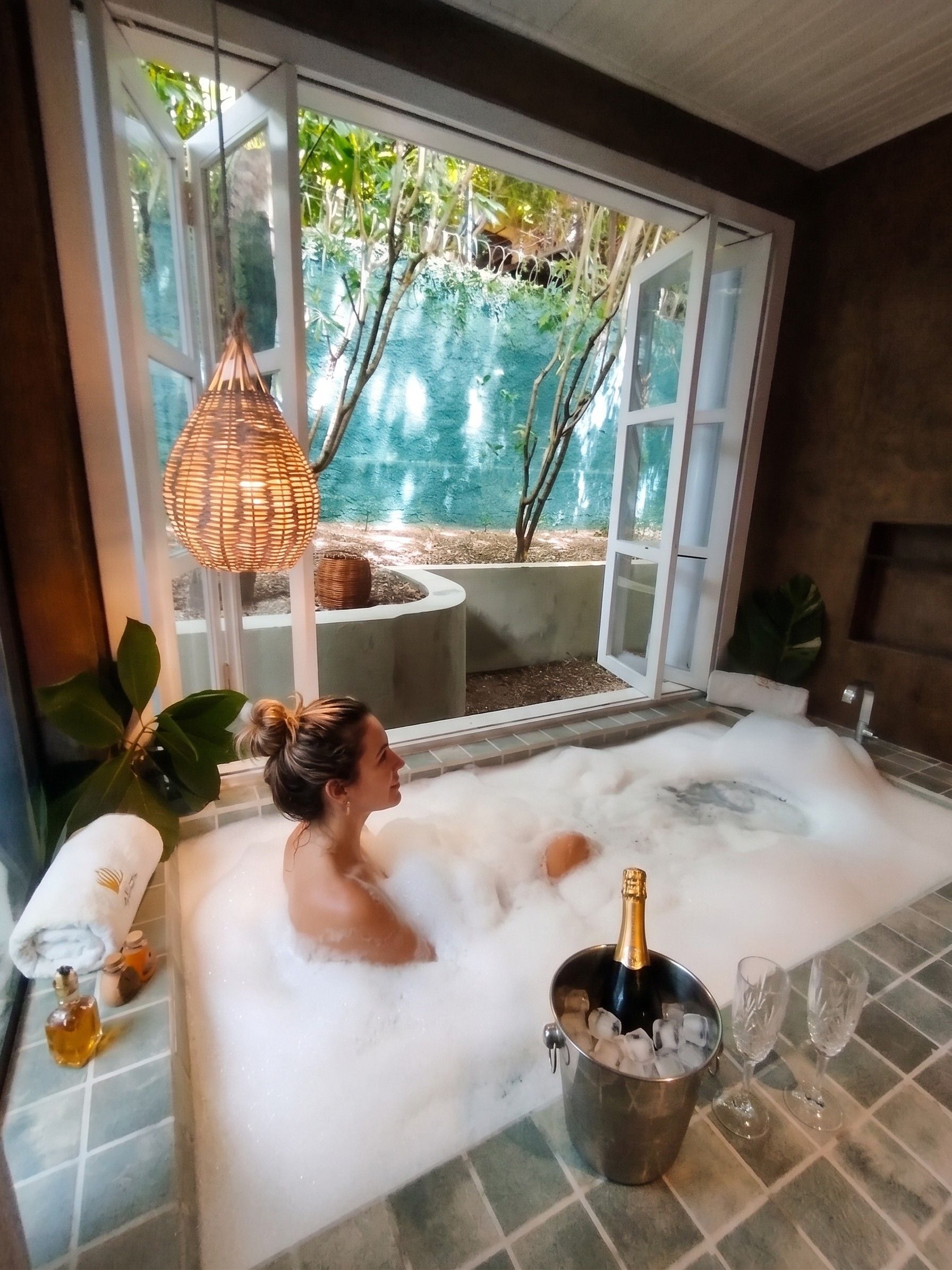 Una persona relajándose en una bañera grande llena de burbujas, con una botella de champán y copas al lado.