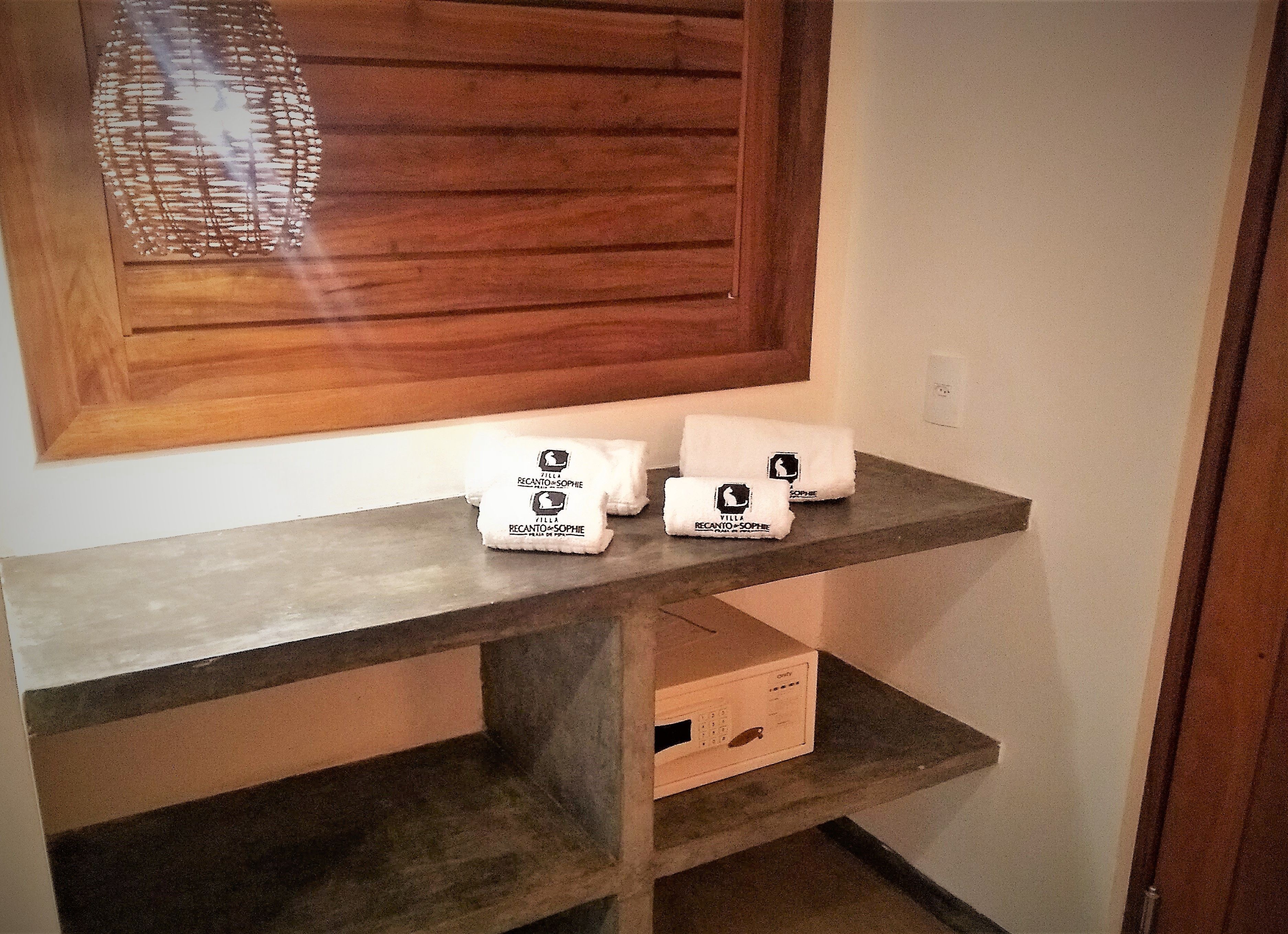 Un estante de madera con tres toallas blancas con la etiqueta "Villa Recanto & Sophie" y una caja pequeña con un control remoto.