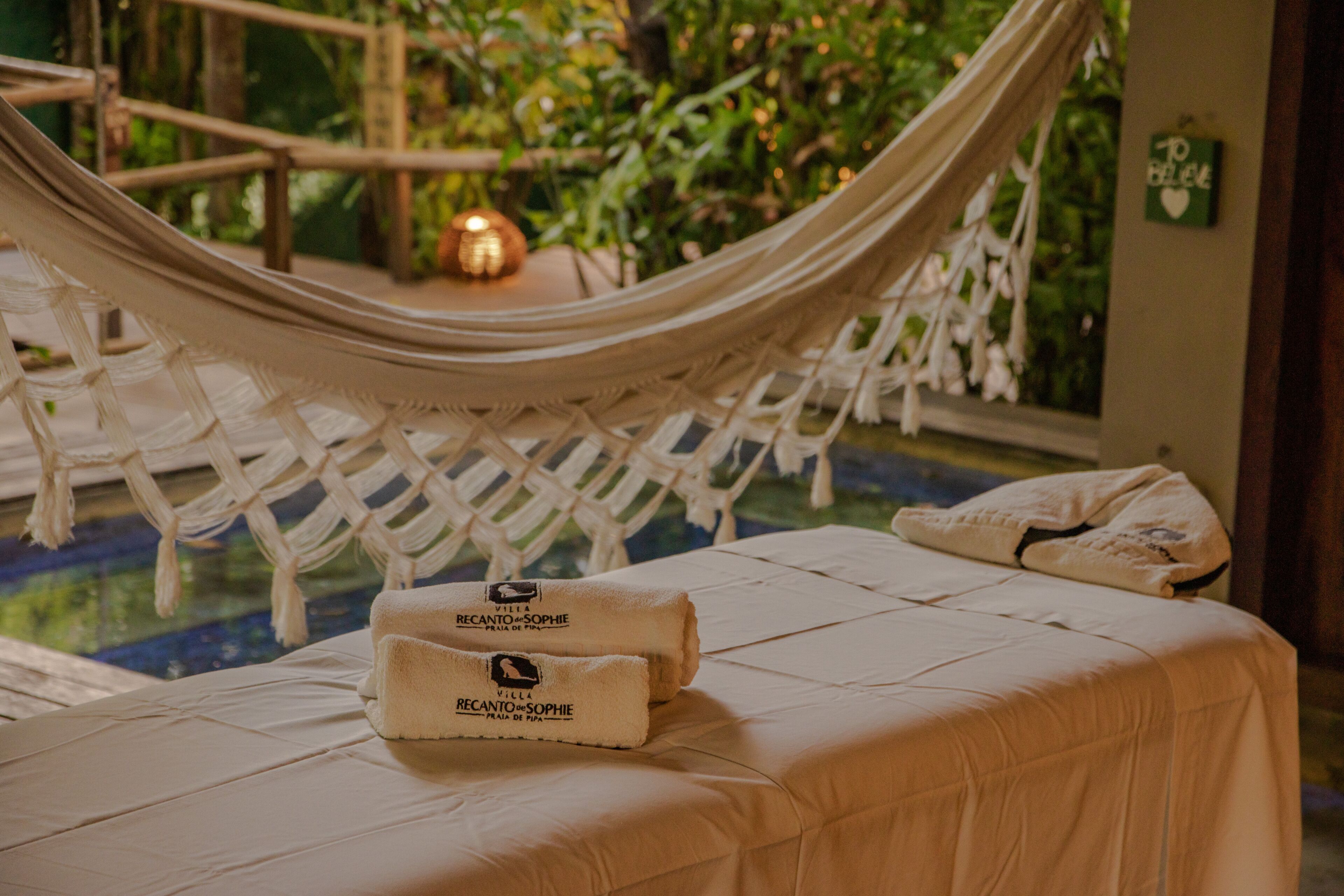 A hammock chair over a bed with towels labeled "Villa Recanto-Sophie" and a small table with a candle.