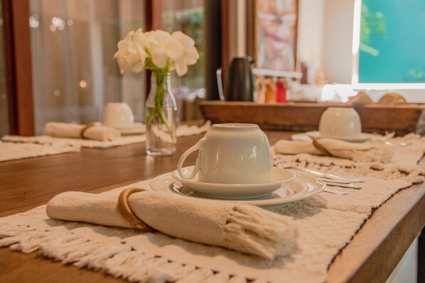 Une table à manger bien mise, avec une tasse et une soucoupe blanches, un vase de fleurs blanches et un plateau en bois sur lequel sont pliées des serviettes.