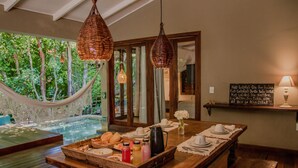Une table de salle à manger en bois, sur laquelle se trouve un plateau avec de la nourriture et des boissons, un hamac et une vue sur un jardin luxuriant, visible à travers de grandes fenêtres.