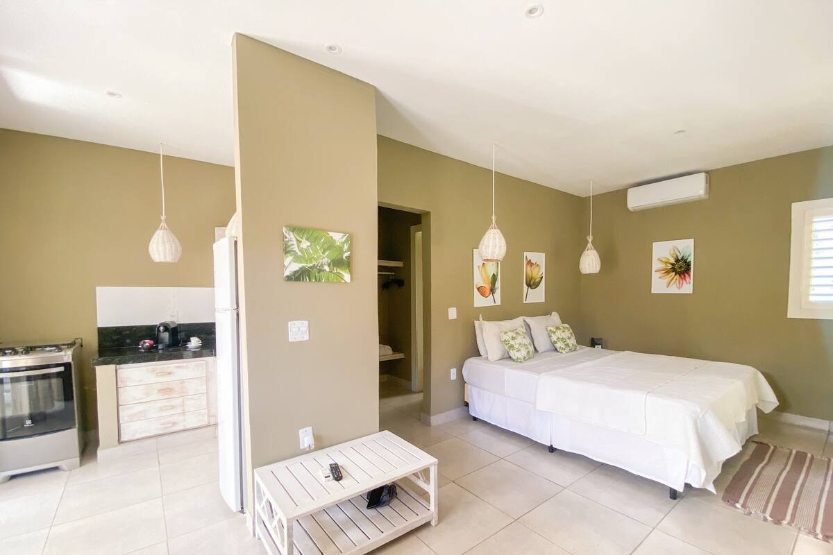 Une chambre moderne avec un grand lit, une table de chevet blanche et un petit banc blanc. Elle comprend un coin cuisine avec une cuisinière et un réfrigérateur. Les murs de la pièce sont de couleur beige et elle possède une grande fenêtre avec des stores.