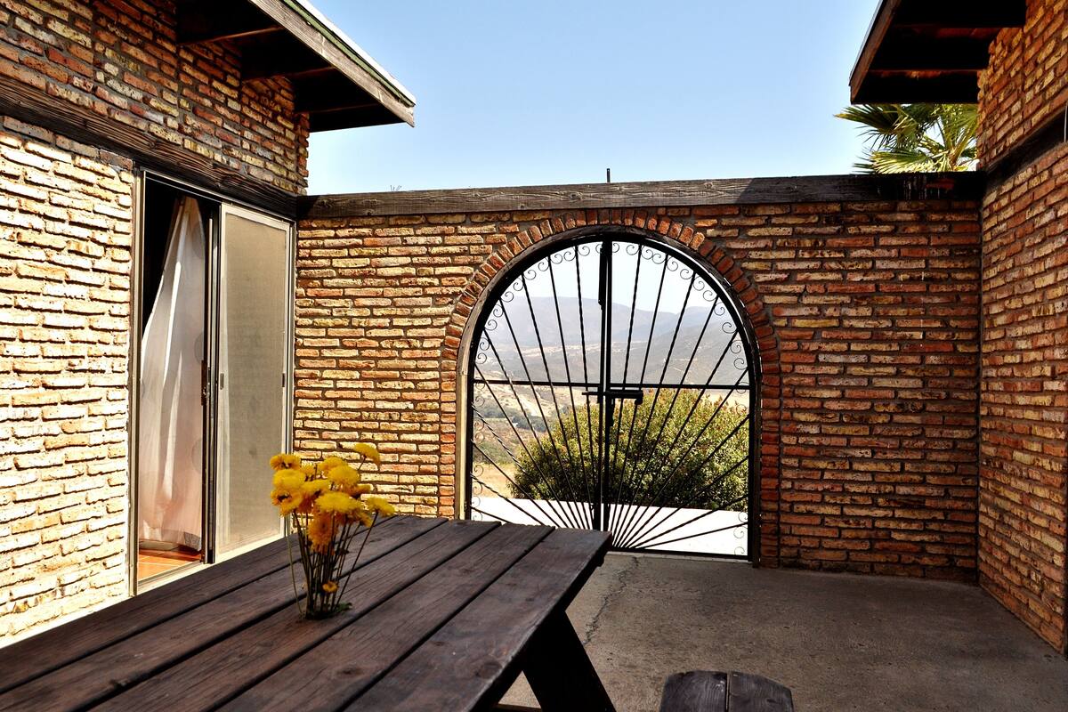 A patio with a wooden table and a vase of yellow flowers, a brick wall, and a large arched window with a view of greenery.