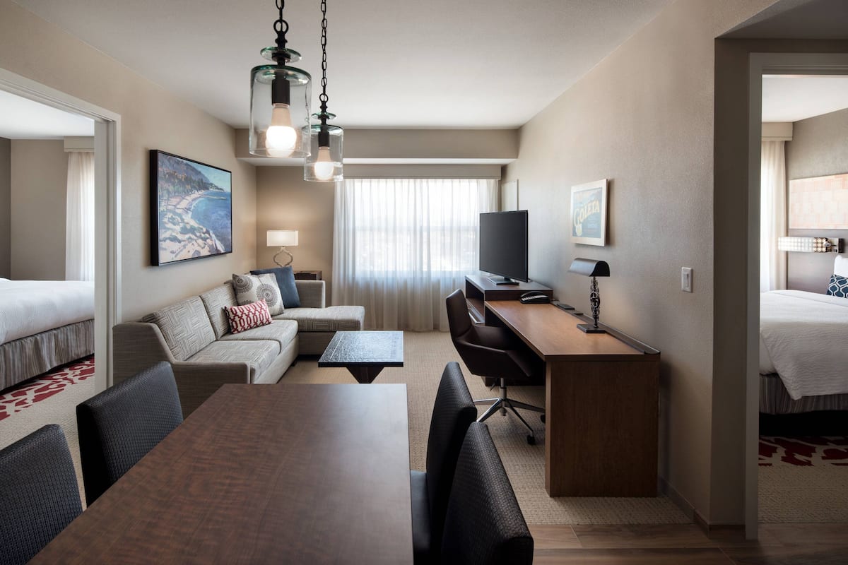 A hotel room with a bed, a sofa, a desk, and a dining table. There is a TV on the desk and a window with curtains.