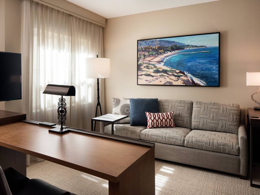 A hotel room with a large flat-screen TV, a wooden desk, a sofa with patterned cushions, a painting of a beach scene, and a lamp on a bedside table.
