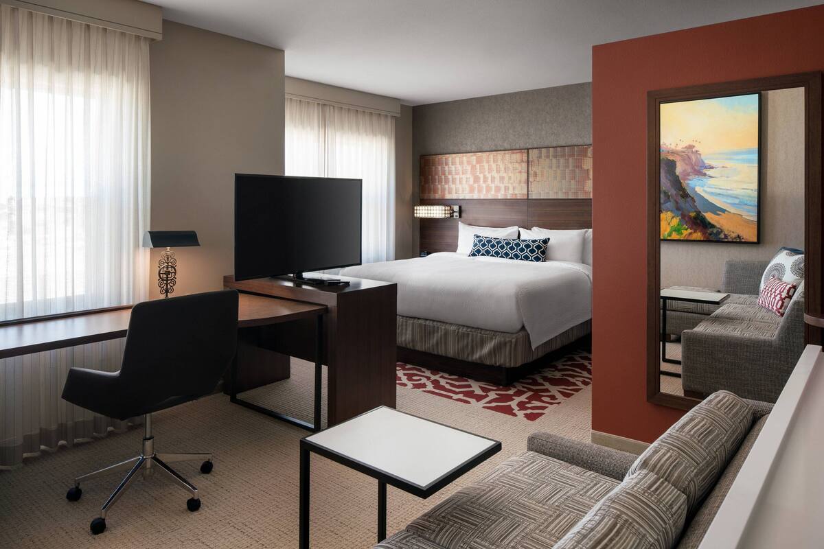 A hotel room with a bed, a desk, a chair, a sofa, a TV, and a painting on the wall.