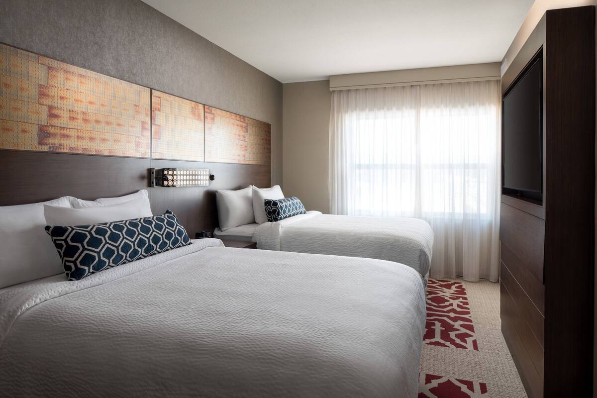 A hotel room with two beds, a large artwork on the wall, a television, and a view of the outdoors through the window.