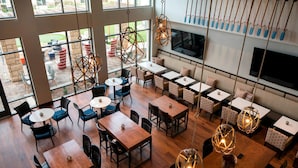 Un restaurant doté de plusieurs espaces de restauration, notamment des tables rondes et des banquettes, agrémenté de luminaires suspendus et d’une grande fenêtre donnant sur l’espace extérieur.