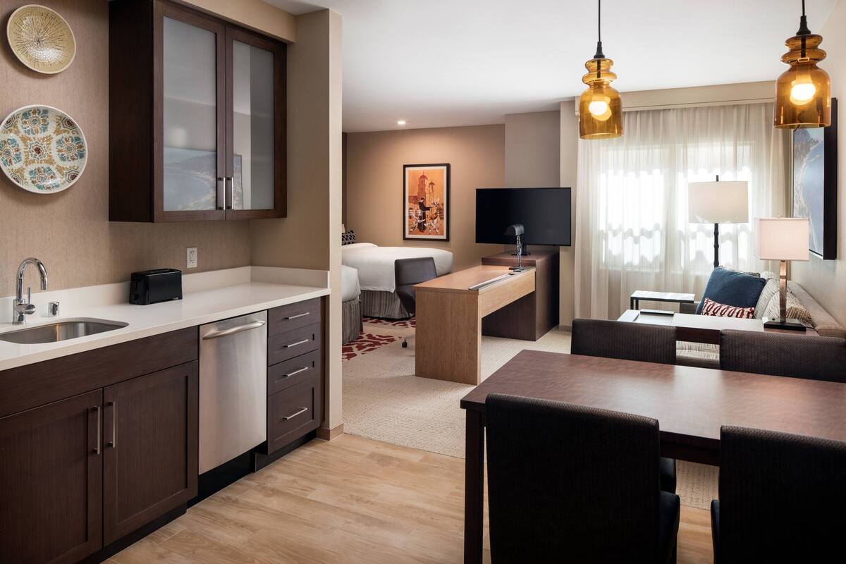 A modern hotel room with a kitchenette, a dining area, a sofa, and a TV.