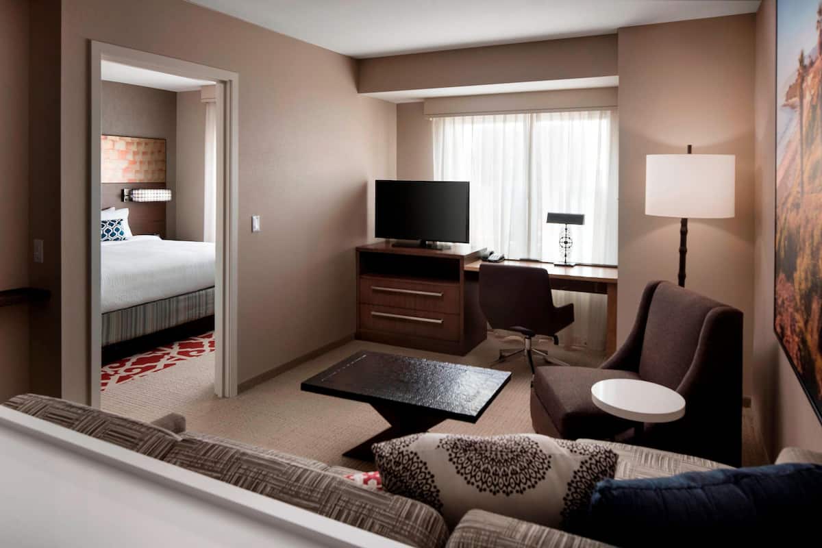 A hotel room with a bed, a desk with a TV, a chair, a sofa, and a lamp.