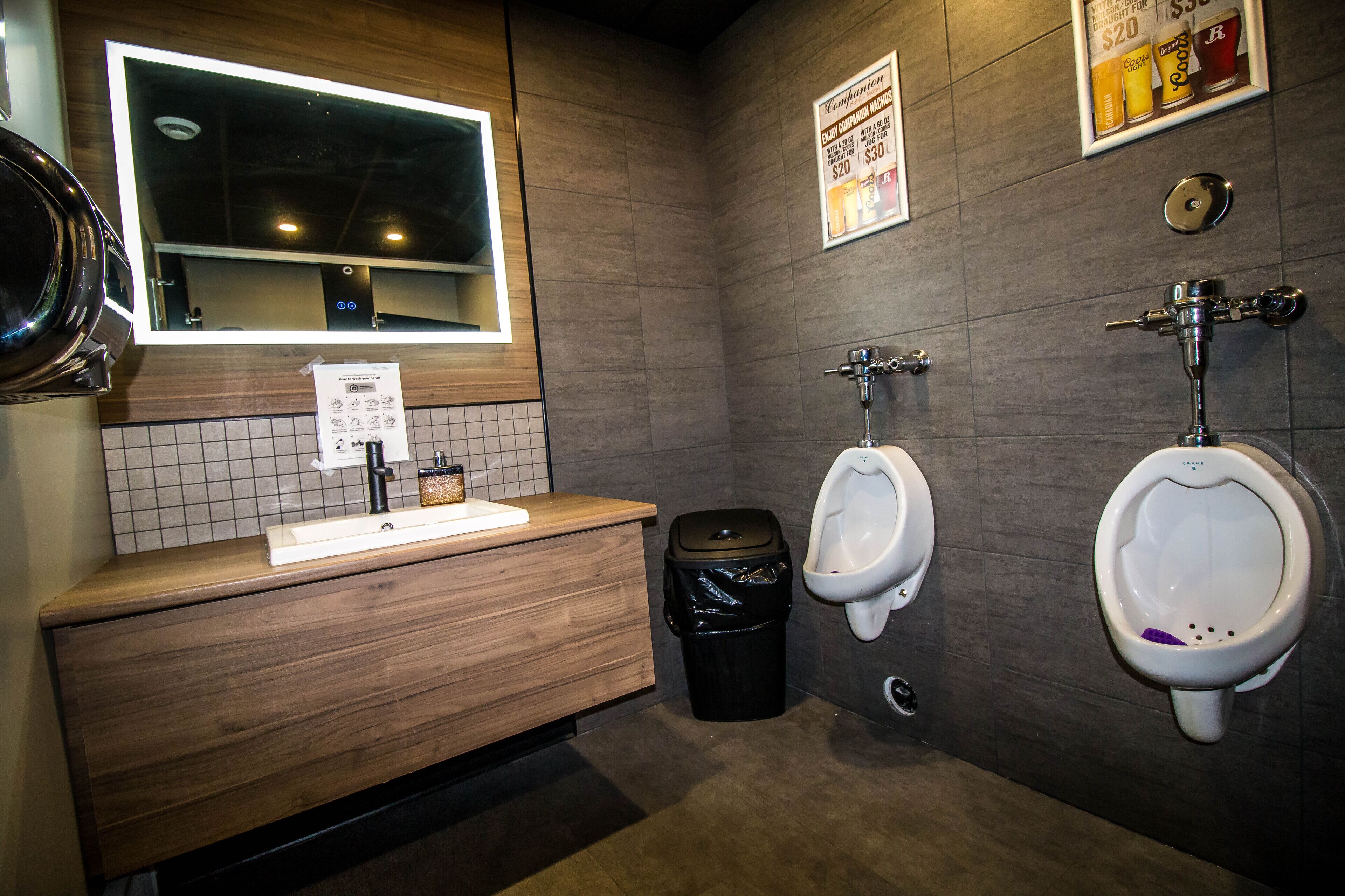 Een herentoilet met een houten wastafelmeubel, een spiegel, twee urinoirs, een prullenbak en een wandbord.