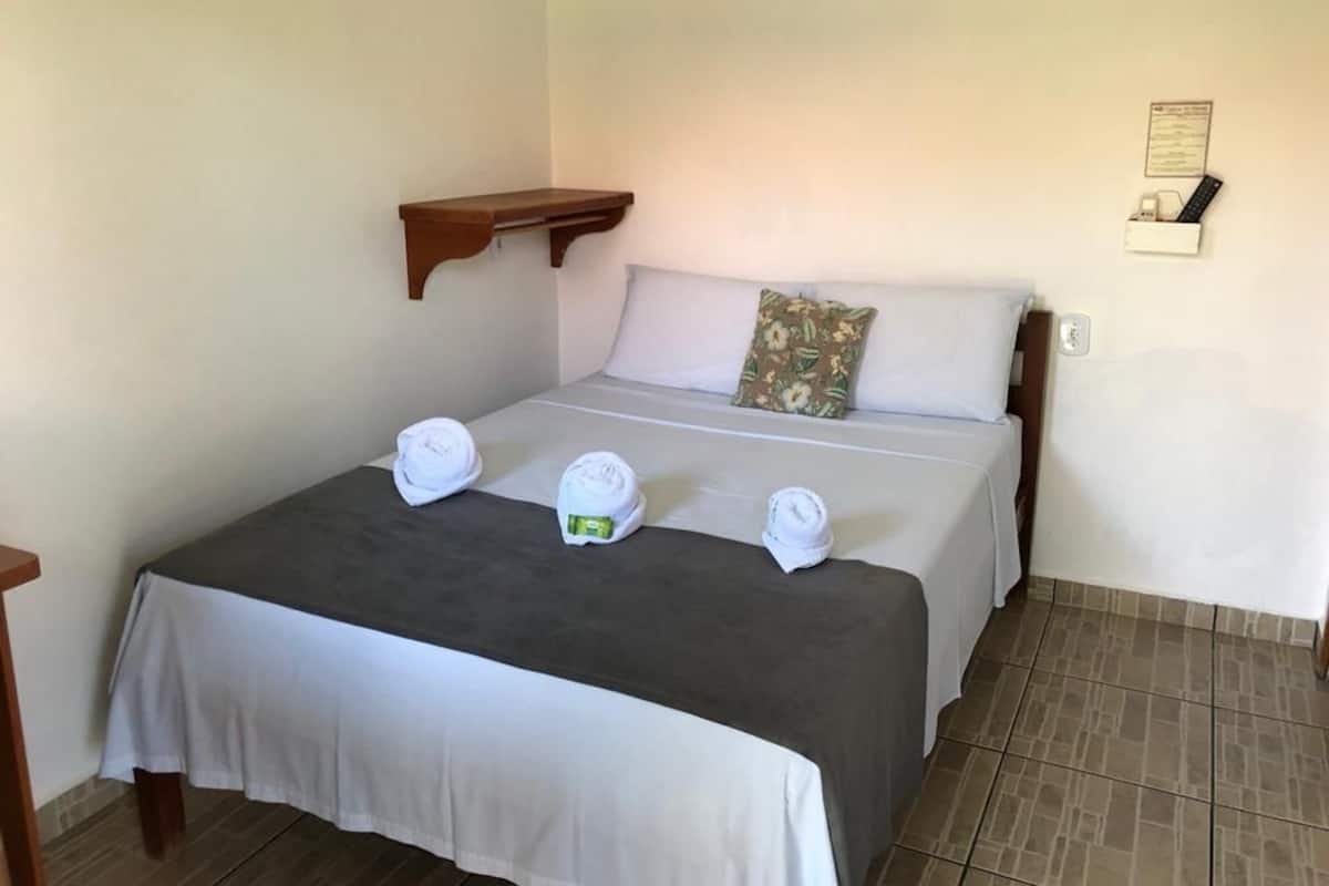 Una cama bien hecha con sábanas blancas y una colcha gris, un estante de madera encima y un objeto pequeño sujeto a la pared a la derecha.