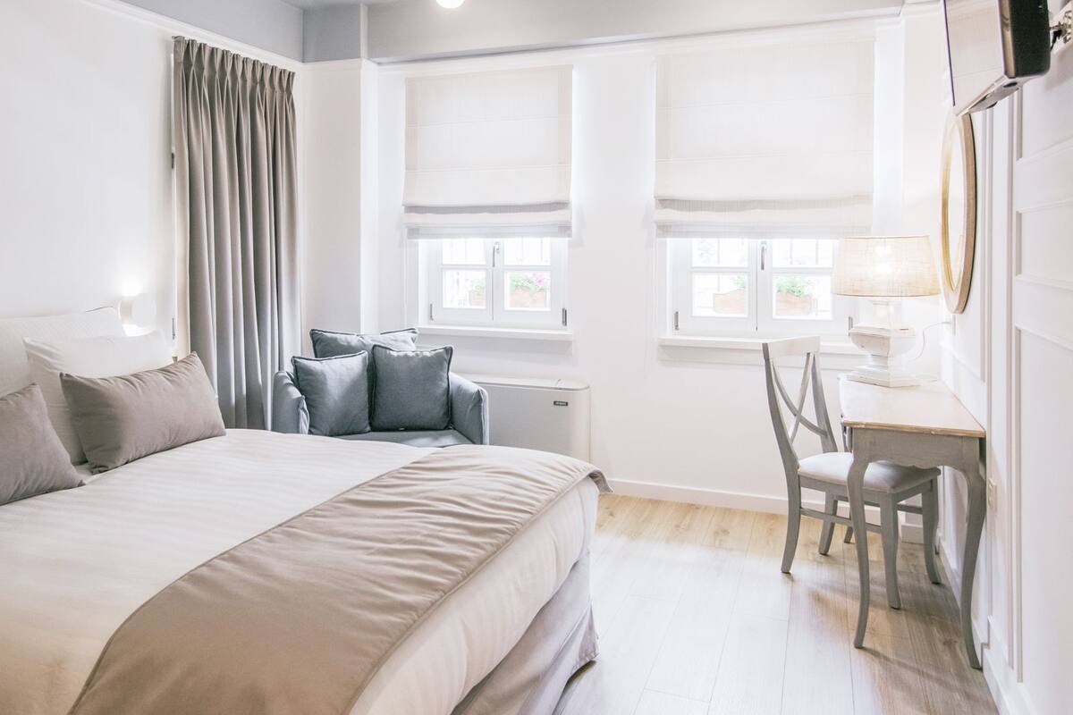 Un dormitorio con una cama grande, un escritorio y una silla. Hay cortinas en la ventana y una lámpara sobre el escritorio.