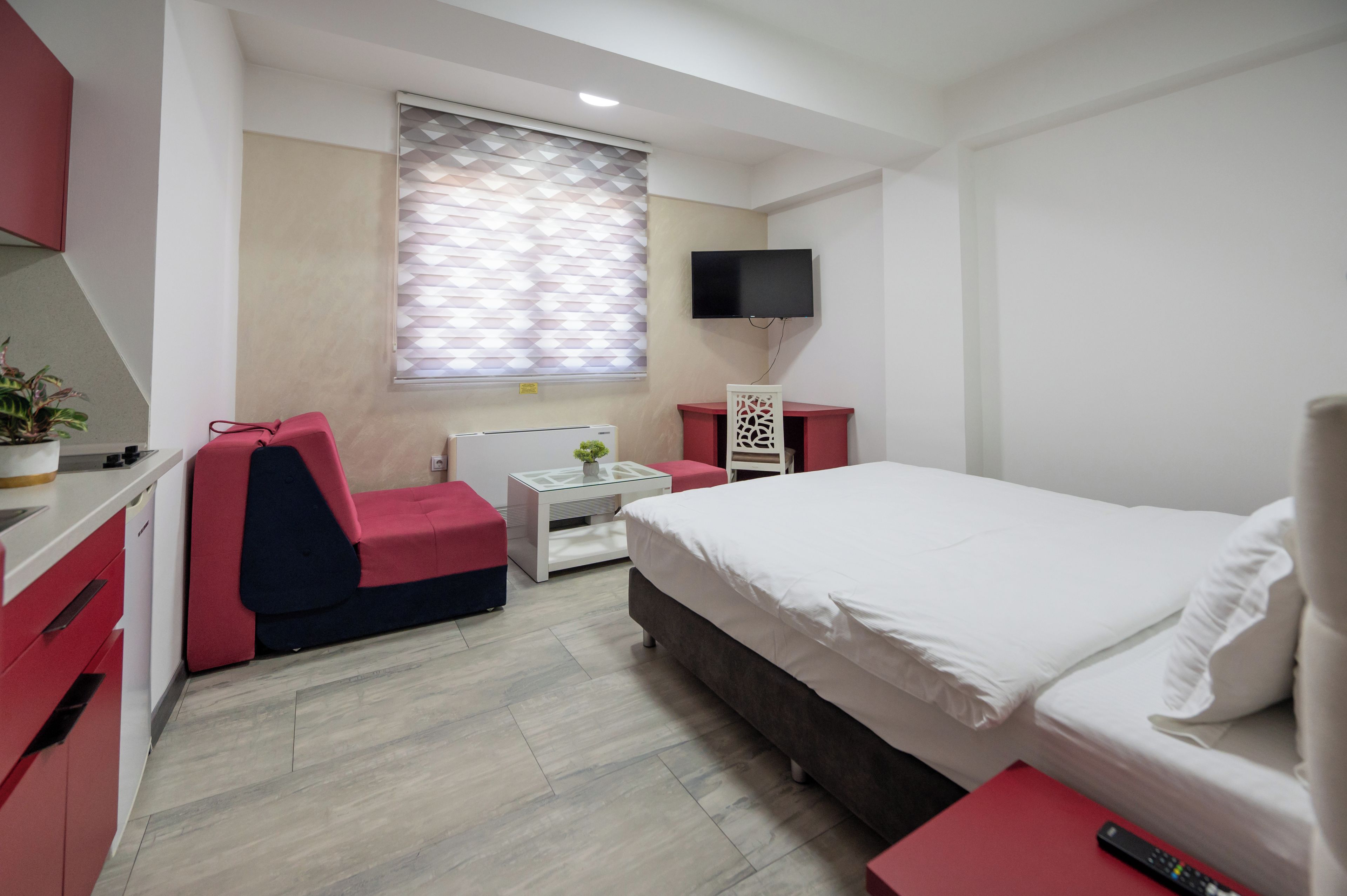 Une chambre d’hôtel comprenant un lit, une télévision, un bureau avec une chaise, une petite table avec une plante et une fenêtre avec des stores.