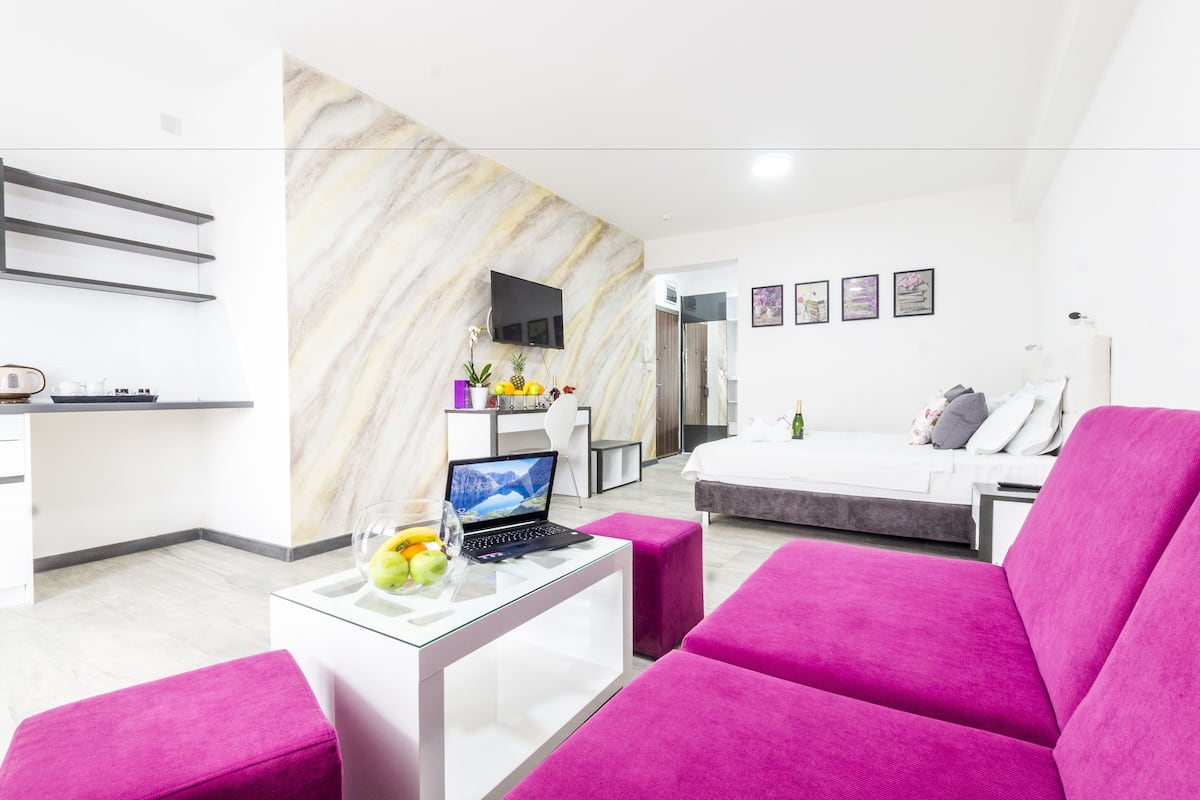 A modern hotel room with a bed, a desk with a laptop, a TV, and a seating area with a pink sofa.