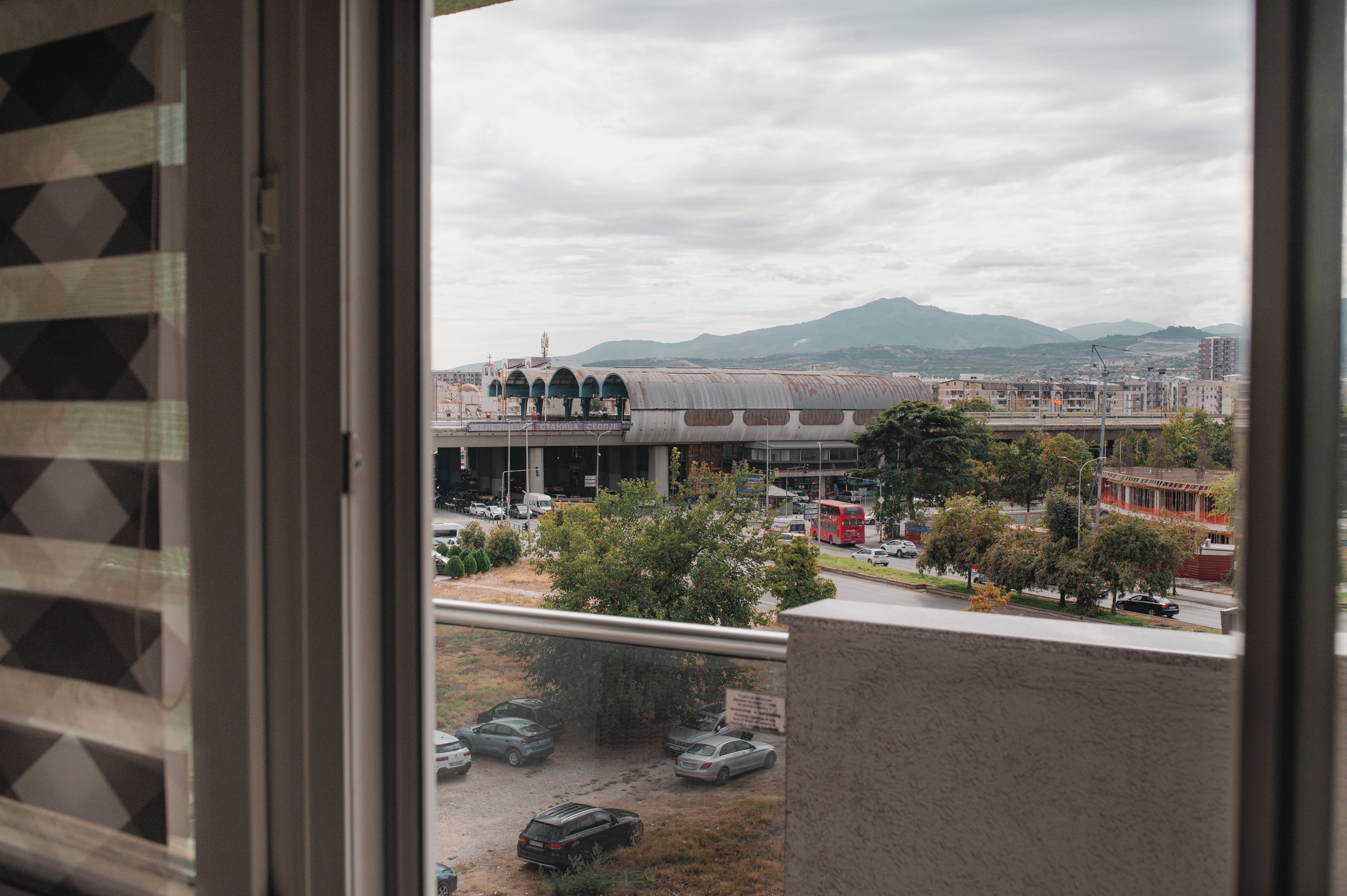 Vista da una finestra su un paesaggio urbano con un grande edificio con cupola e strutture ad arco, un'autostrada con veicoli e montagne in lontananza.