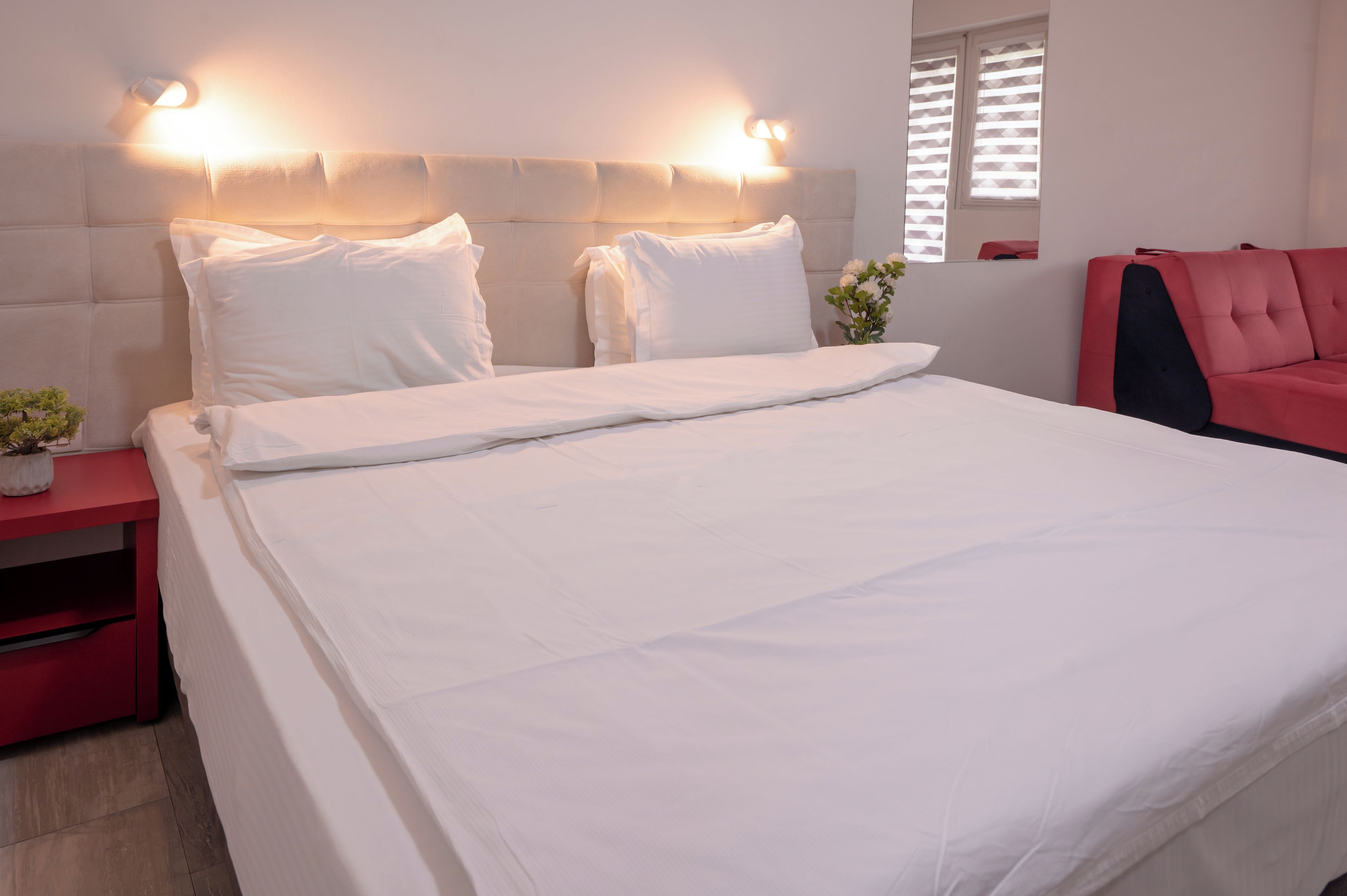A neatly made bed with white linens, a red nightstand with a small plant, and a red chair to the right.