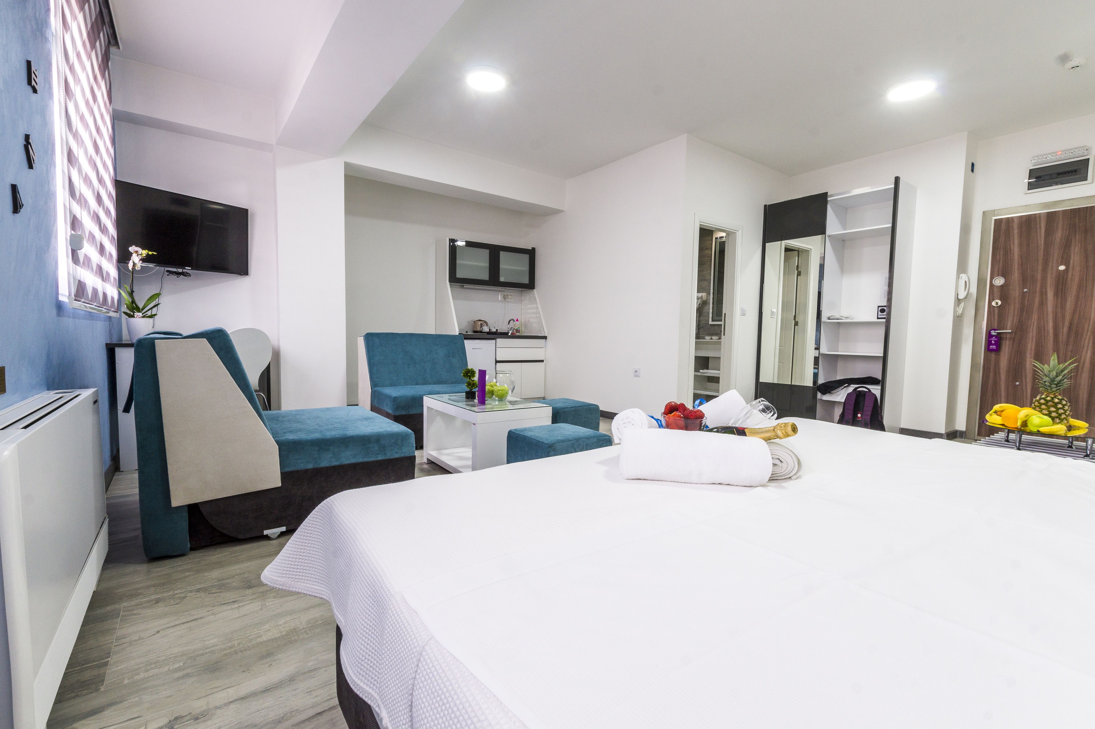 Une chambre d’hôtel comprenant un lit, un coin salon avec un fauteuil et une petite table, une kitchenette, un téléviseur fixé au mur et une armoire.