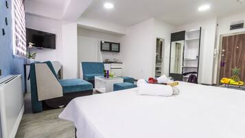 Une chambre d’hôtel comprenant un lit, un coin salon avec un fauteuil et une petite table, une kitchenette, un téléviseur fixé au mur et une armoire.