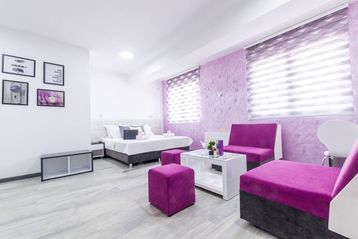 A modern hotel room with a white bed, purple and white furniture, and a large window with blinds.