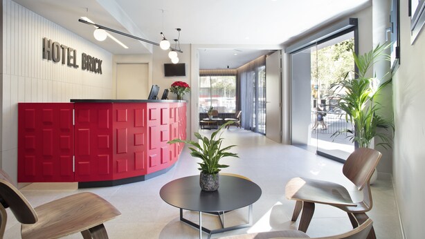 Een moderne hotel receptie met een rode balie, een ronde tafel met stoelen en een groot raam met uitzicht op de omgeving.