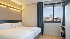 Een hotelkamer met twee bedden, een bureau en een groot raam met uitzicht op de stad.
