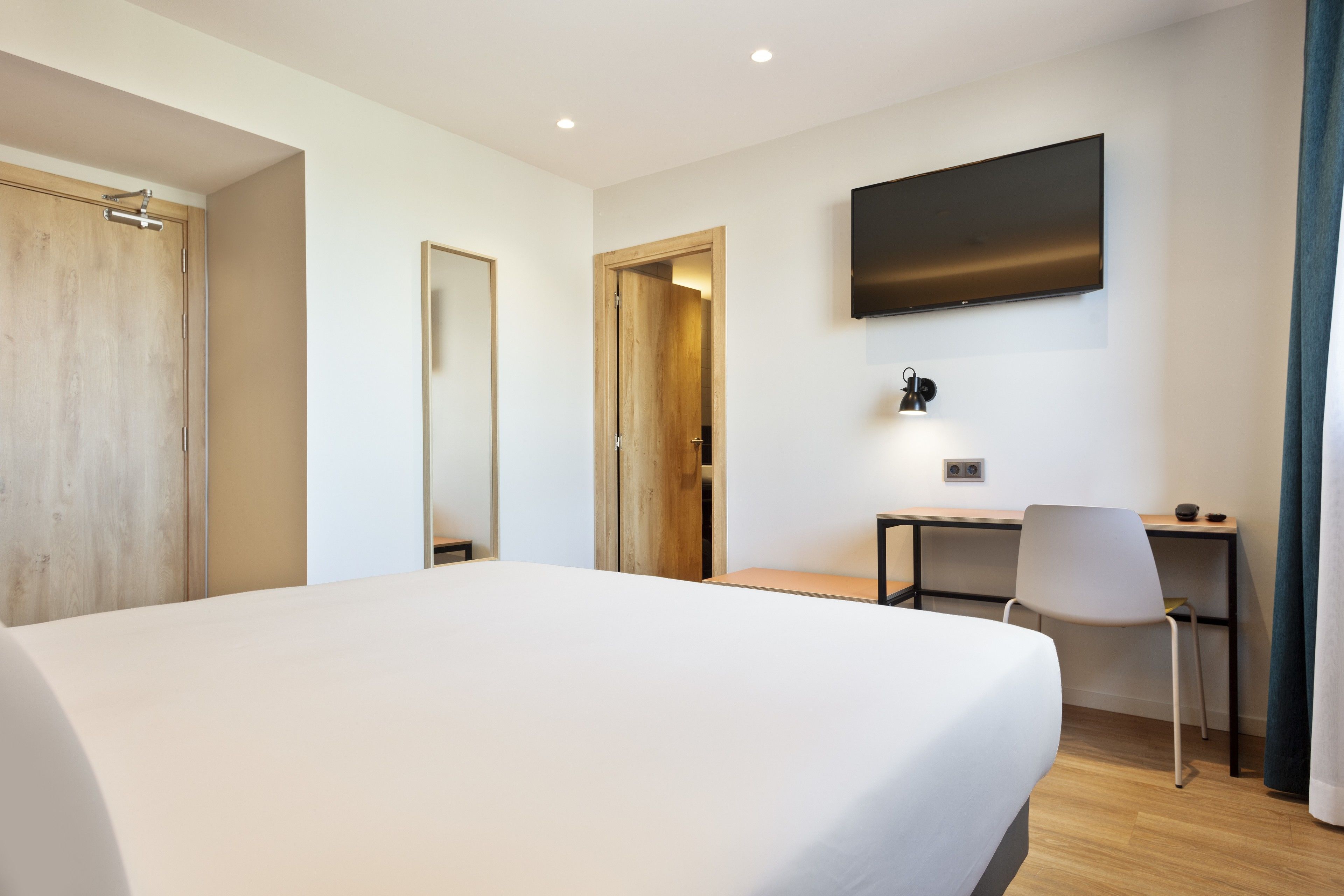 Een hotelkamer met een groot bed, een bureau, een stoel, een aan de muur bevestigde televisie en een deur die naar een andere kamer leidt.